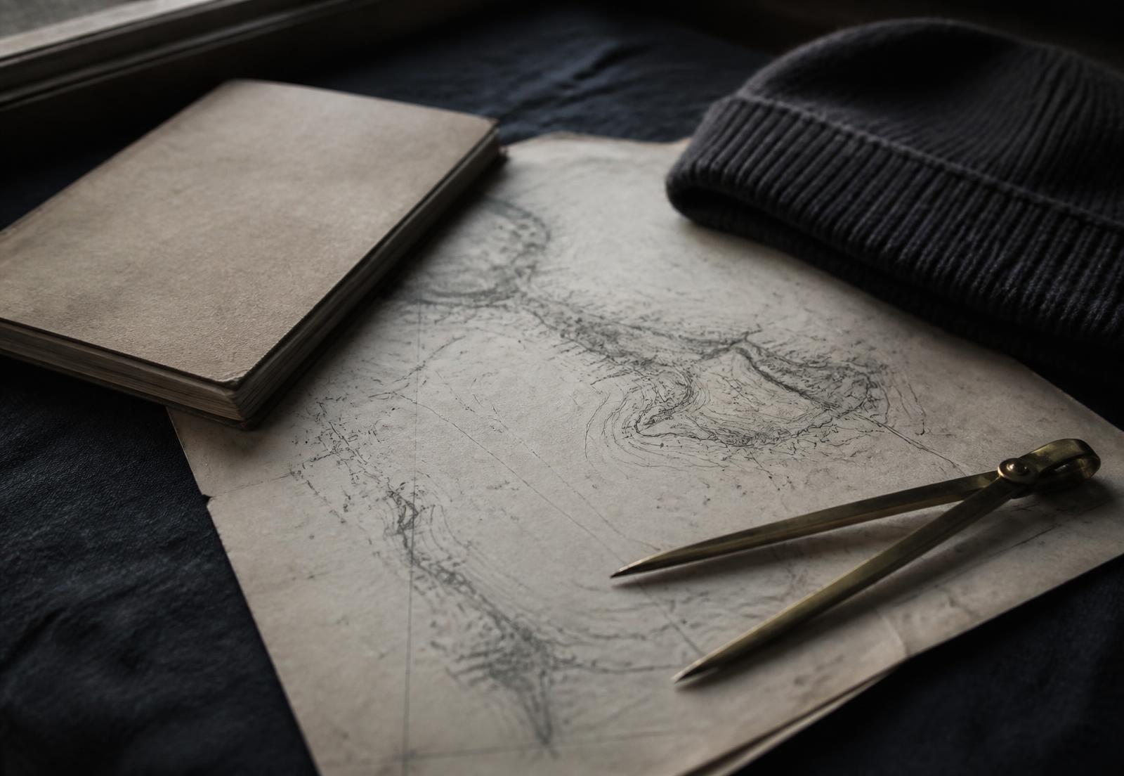 Chart paper marked with pencilled contour lines beside a notebook, dividers and a wool cap